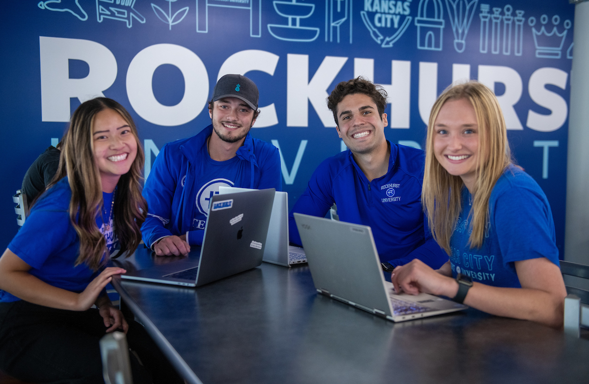 Four students in Rockhurst blue with laptops in front of Rockhurst mural
