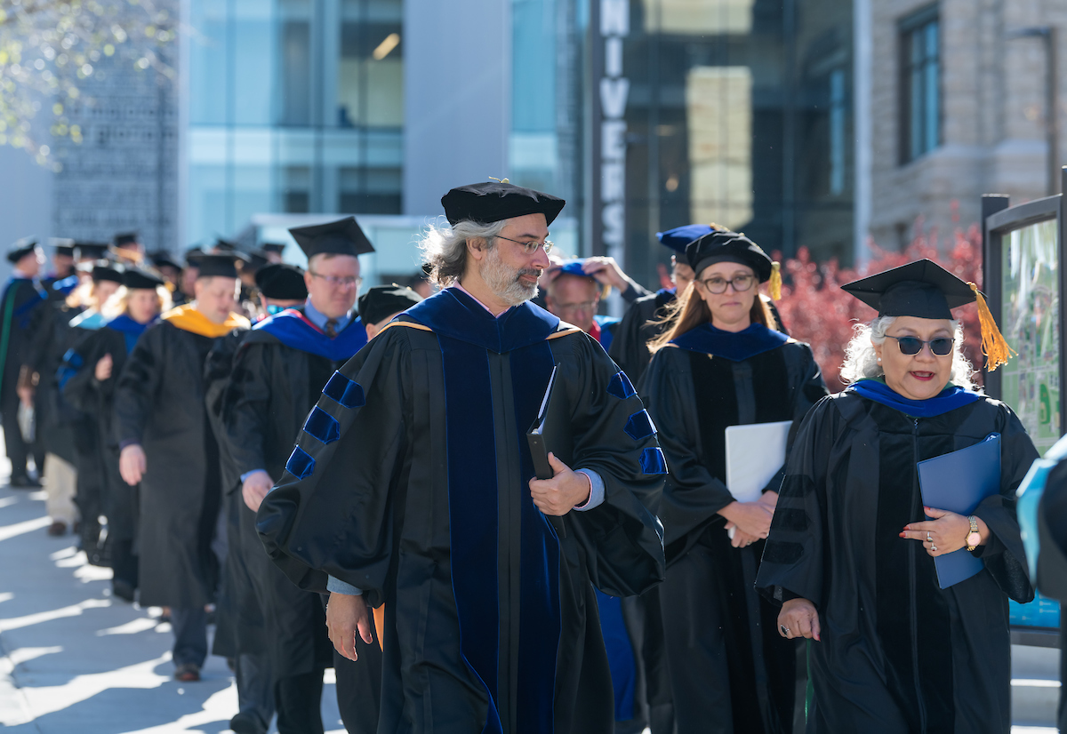 Faculty in full commencement attire walk in a line between buildings