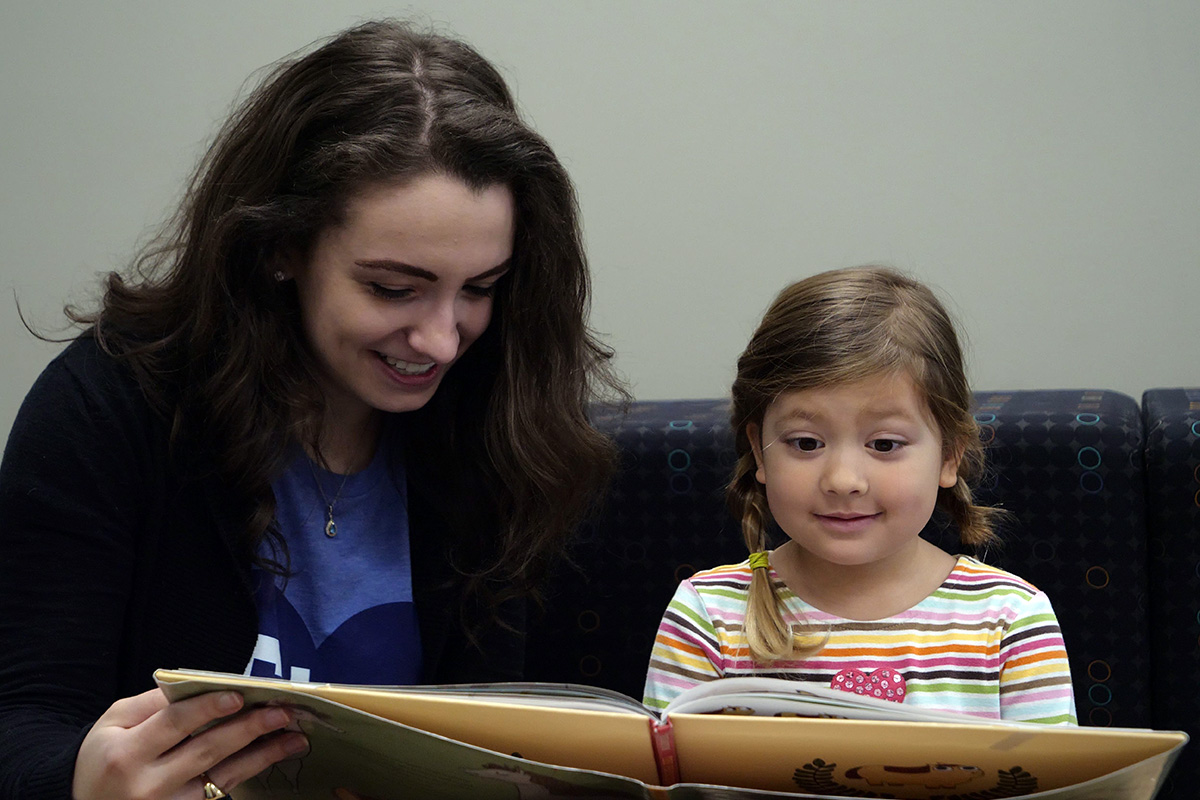 A female SLP student touches reads a book with a smiling child client