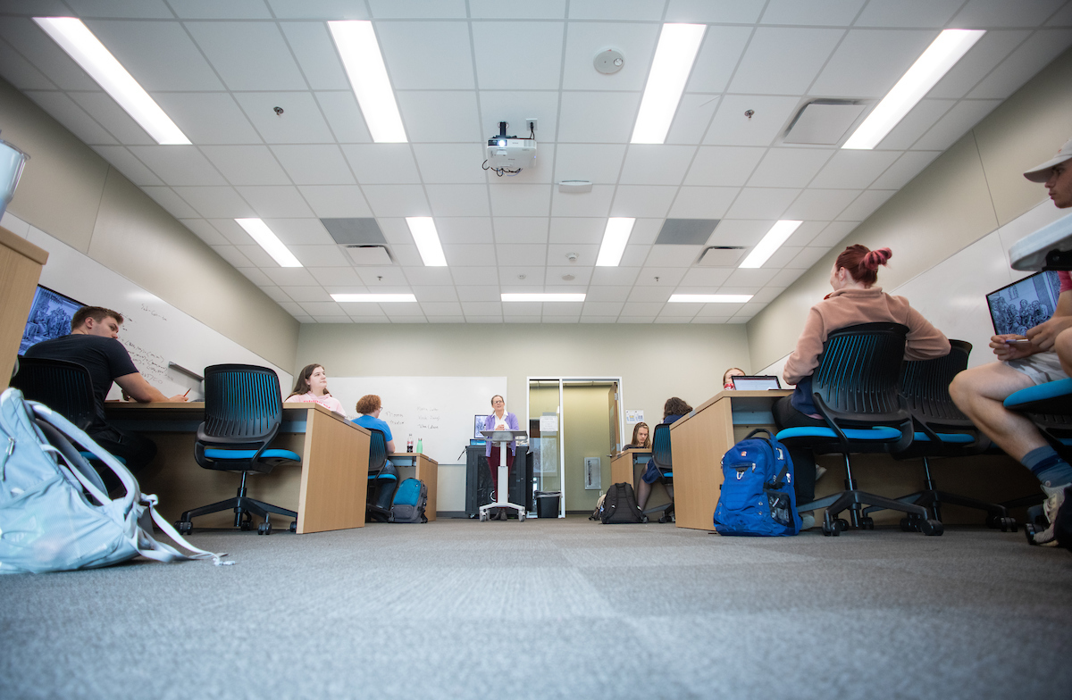 View of a classroom from the floor in the back. A teacher is visible at the front of the room behind a podium. 