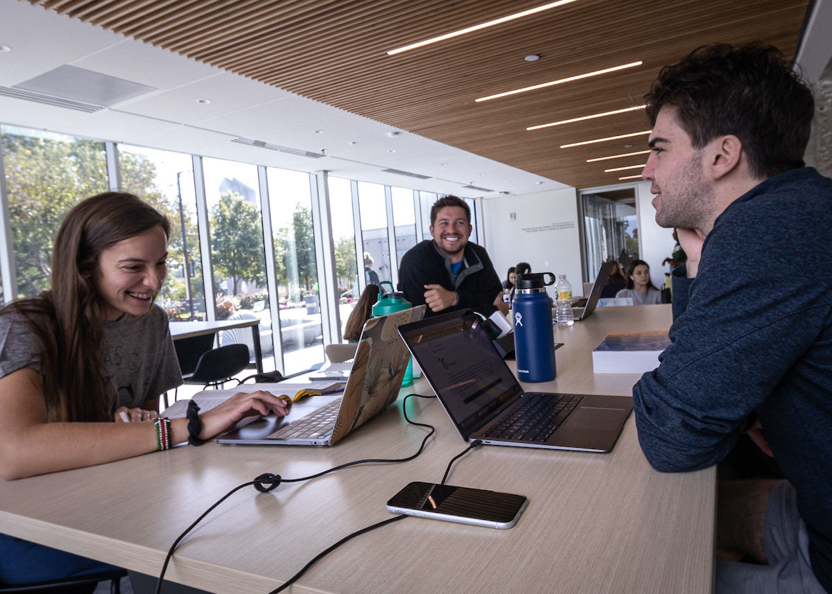 A room full of accounting students working. One student is smiling and looking at the camera. Two other students in the foreground of the picture discuss their CPA Pathway Accounting certificate. 