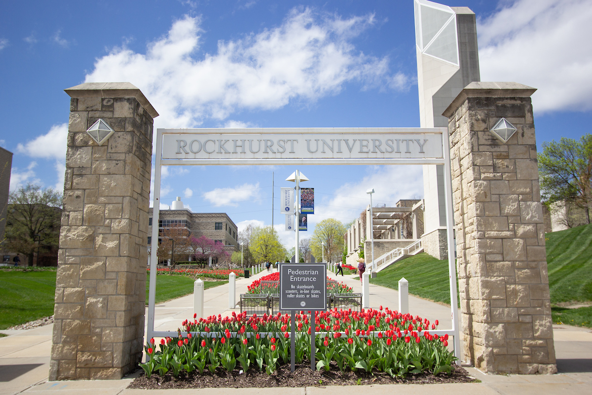 A Rockhurst University sign over a pedestrian entrance with tulips underneath.