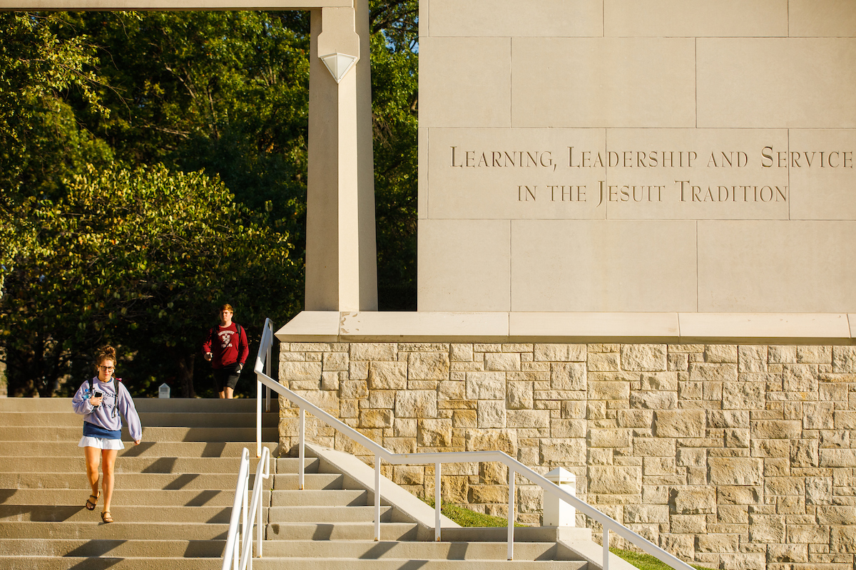 An inscription of "Learning, Leadership and Service in the Jesuit Tradition" is seen on a bell tower as students take the adjacent stairs.