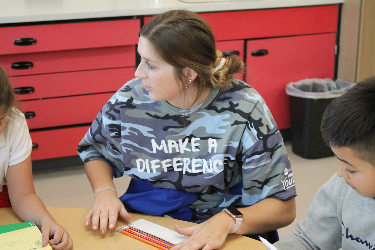 A student teacher talks with kids while seated at a table