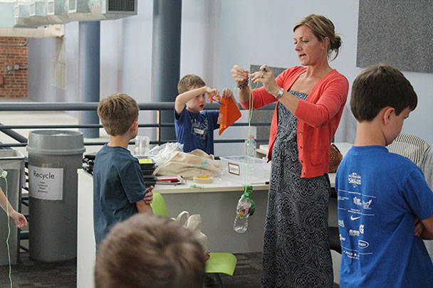 A woman leads an elementary school class in a science experiment with a plastic bottle on a string