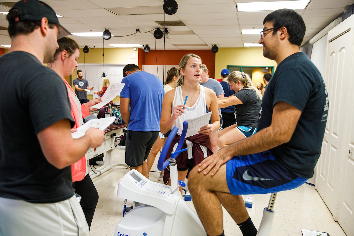 A volunteer sitting on an exercise bike takes questions from PT students