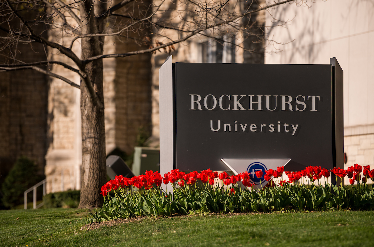 A Rockhurst University sign sits in front of campus, surrounded by red tulips with trees in the background. 