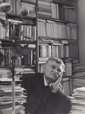 Gabriel Marcel sits at his desk, surrounded by piles of books 