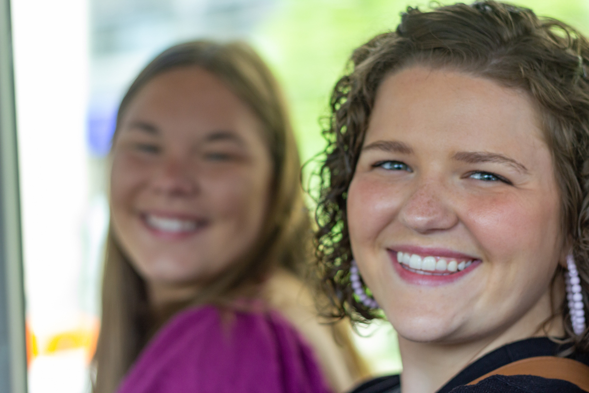 A student in the foreground, in focus, smiles at the camera. Another student, out of focus in the background, is also smiling. 