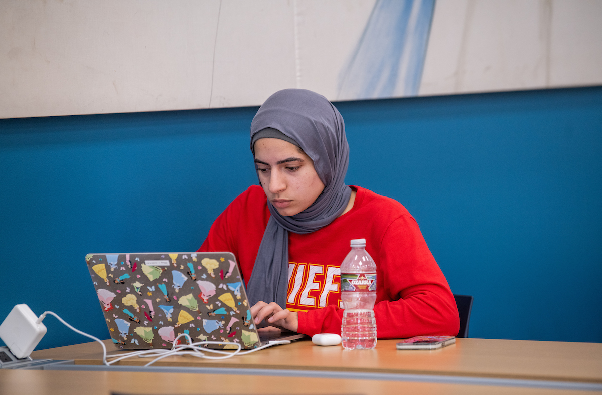 A girl wearing a Chiefs shirt and hijab works on a colorful laptop