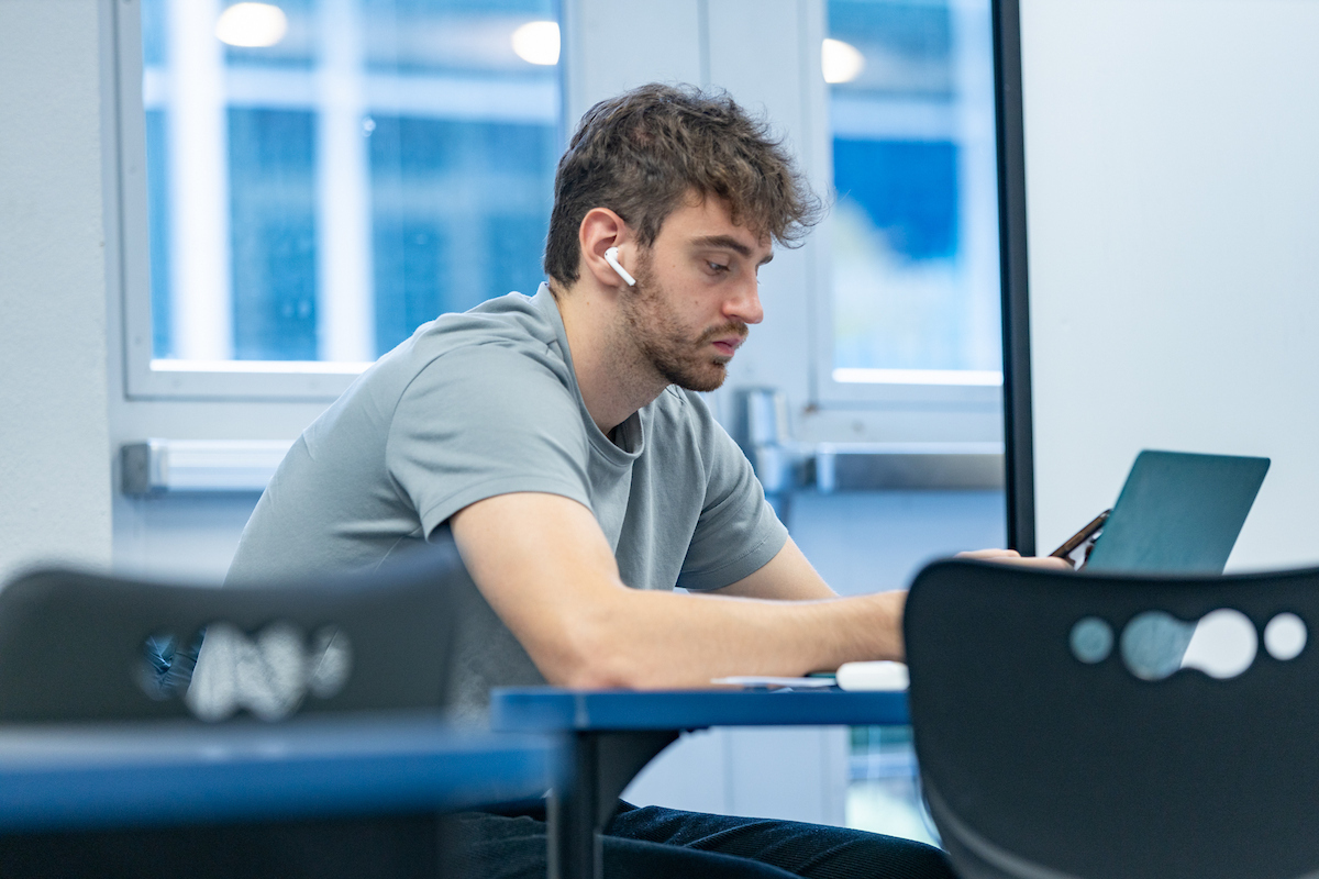 A male student with an air pod in his ear types on a laptop at a table