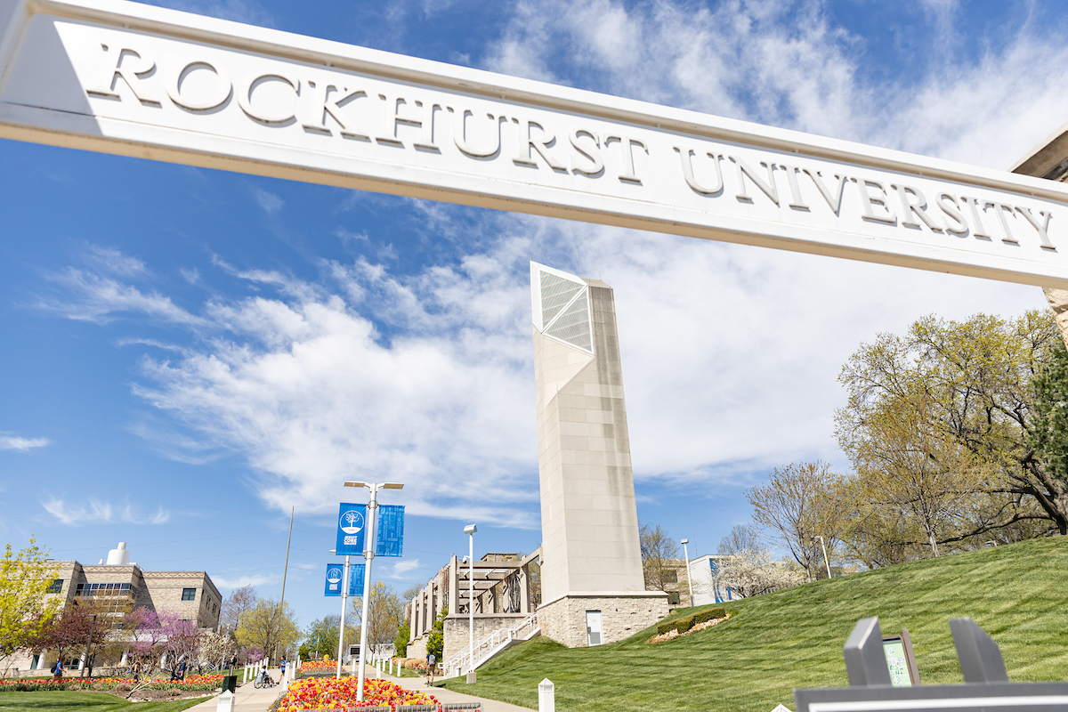 A sign saying Rockhurst University is seen with the campus bell tower and tulips in the background