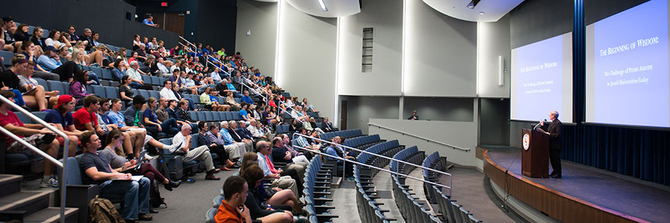 Audience sits in Arrupe Auditorium while speaker stands before two screens