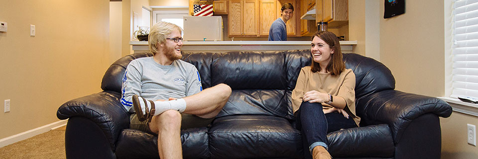 Students sit on a living room couch while another student stands behind them in the kitchen