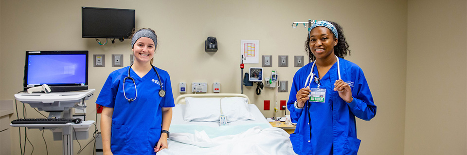 Two nursing students pose beside a practice hospital bed