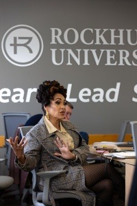 A woman speaks while gesturing with her hands during an EMBA class. Rockhurst University branding is on the wall behind her.