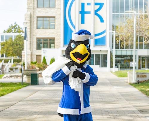 Rock E. dressed as Santa in a blue suit standing in front of Sedgwick Hall with a bag.