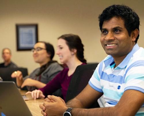 male student smiling while sitting at desk, two female students in background