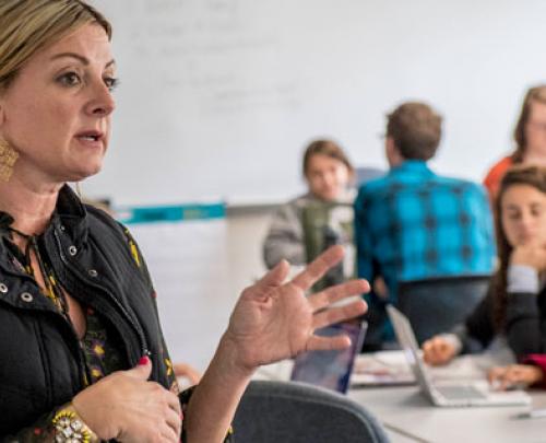 female teacher gesturing with hand in foreground with multiple students in the background