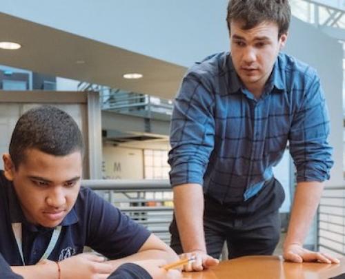 two male students, one sitting, one standing, at a table