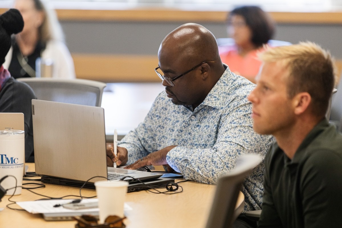 A man with an open laptop writes on a notepad while sitting in at a table in a classroom