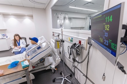 A medical student is writing health data next to a patient in a hospital bed. The picture focuses on the medical equipment on the other side of the bed. 