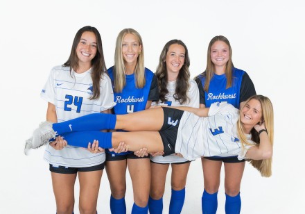 Five women's soccer players pose on media day, the fifth being held horizontal in front of the others. 