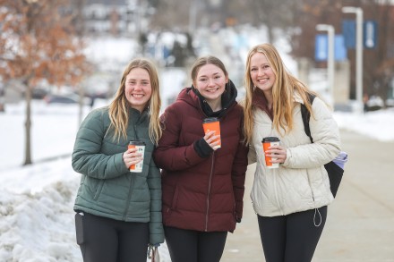 Three friends holding coffee cups pose for the camera with snow behind them