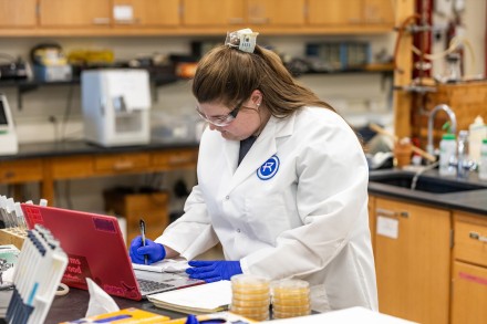 A female student in a Rockhurst lab coat makes notes in a Chemistry lab