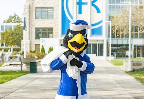 Rock E. dressed as Santa in a blue suit standing in front of Sedgwick Hall with a bag.