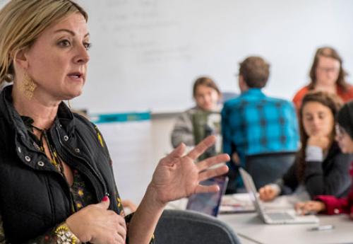 female teacher gesturing with hand in foreground with multiple students in the background